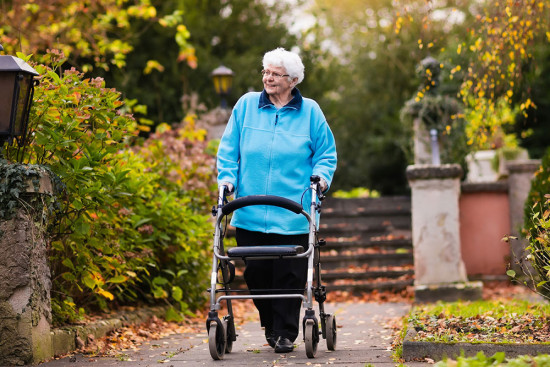 An smiling older woman in a blue top walks through a park using a walking frame with wheels. 